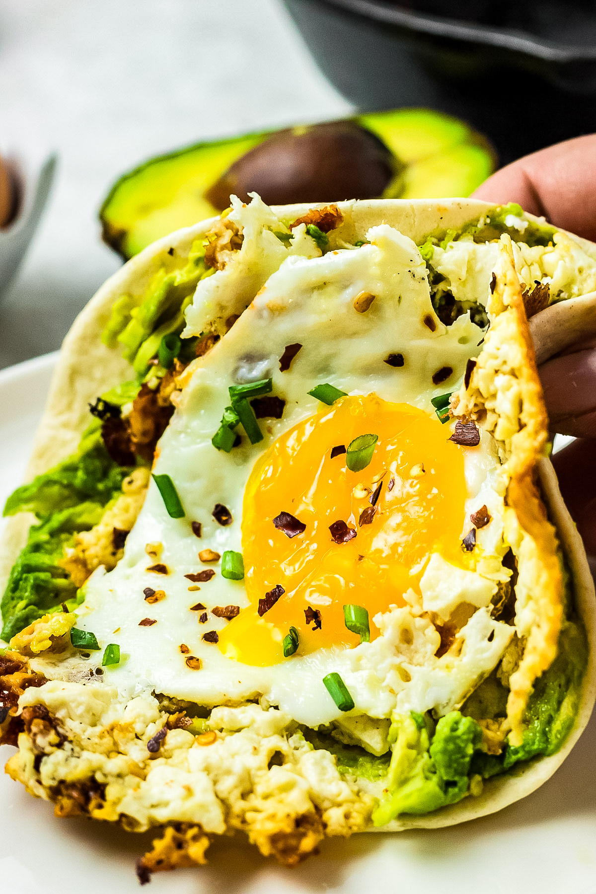 A hand holds a tortilla wrap filled with avocado, a crispy fried egg, and crumbled feta cheese, garnished with chives and red pepper flakes. An avocado is in the background.