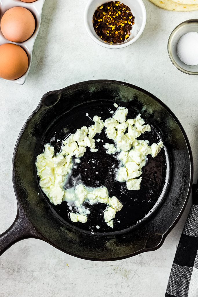 A cast iron skillet on a countertop with crumbled feta cheese in it. Surrounding the skillet are crispy fried eggs, a dish with red pepper flakes, and a small bowl of salt.