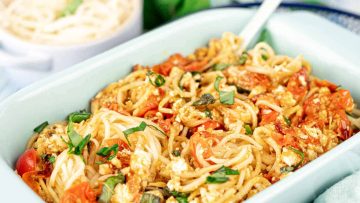 A close-up of a dish featuring pasta mixed with diced tomatoes, basil, and crumbled tofu in a light blue rectangular dish. Another bowl of pasta is partially visible in the background.