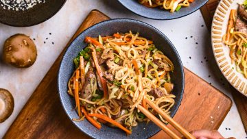 A bowl of stir-fry noodles with vegetables and meat, garnished with black sesame seeds. A hand is holding chopsticks, lifting some noodles from the bowl. Mushrooms are on the table.
