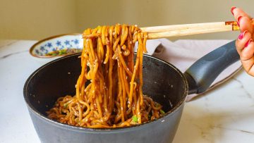 A hand using chopsticks to lift cooked noodles from a black pot on a marble countertop. The noodles are coated in a brown sauce and sprinkled with green onions.