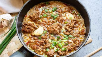 A pan of spicy noodle soup with ground meat, green onions, and garlic cloves on a woven mat.