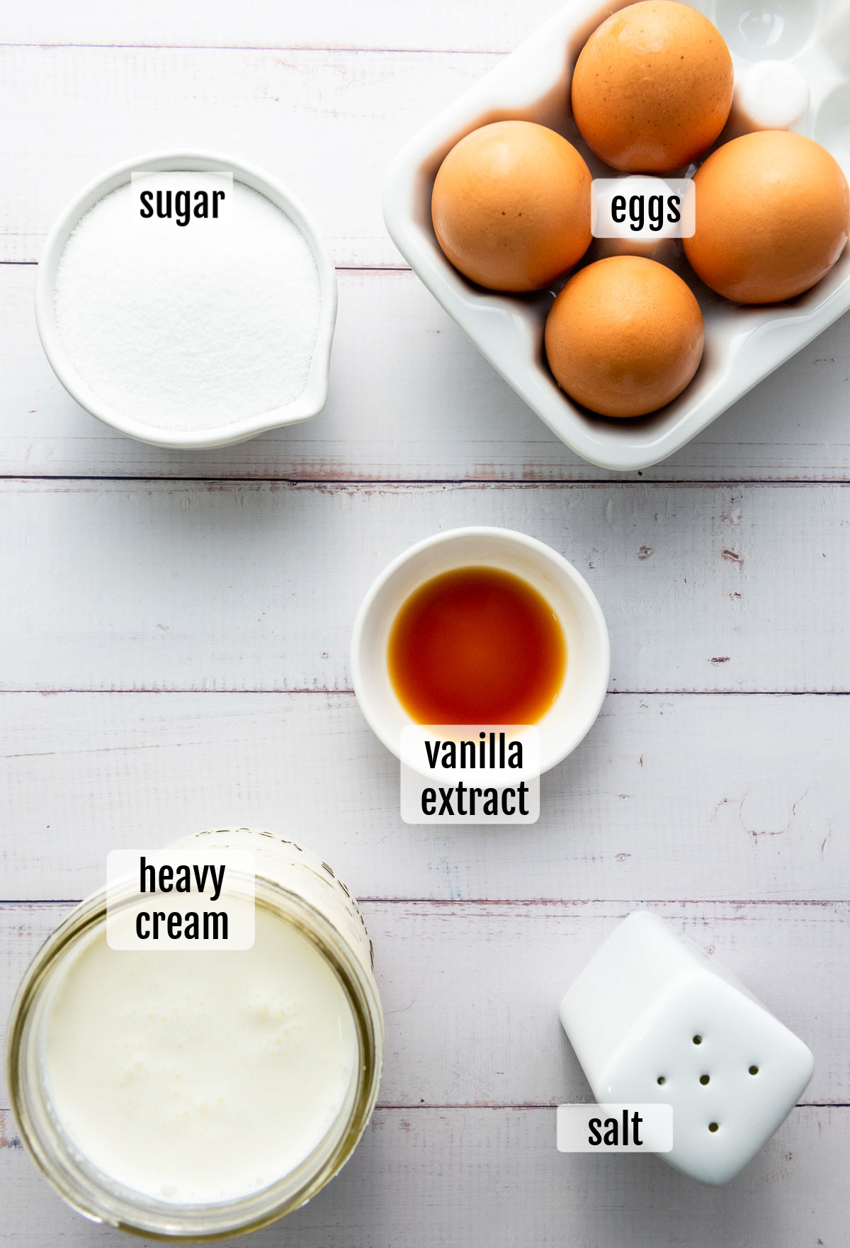Ingredients for Crème Brûlée are beautifully arranged on a table: a bowl of eggs, a container of sugar, a cup of heavy cream, a small bowl of vanilla extract, and a salt shaker.