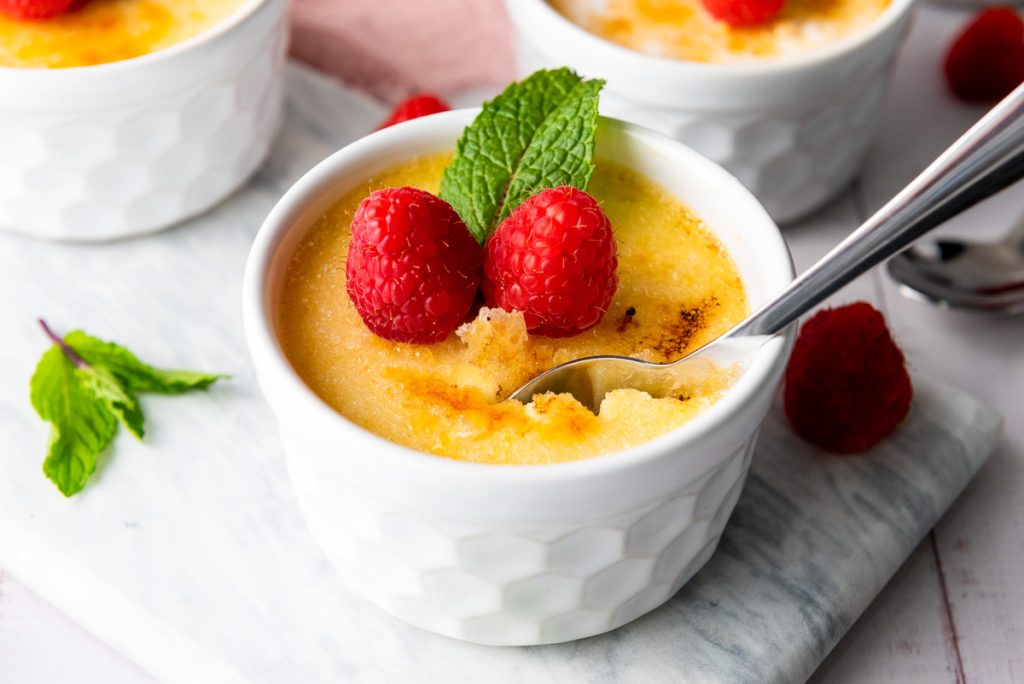 A ramekin of sumptuous Crème Brûlée crowned with two ripe raspberries and a mint leaf, invitingly paired with a spoon nestled inside.