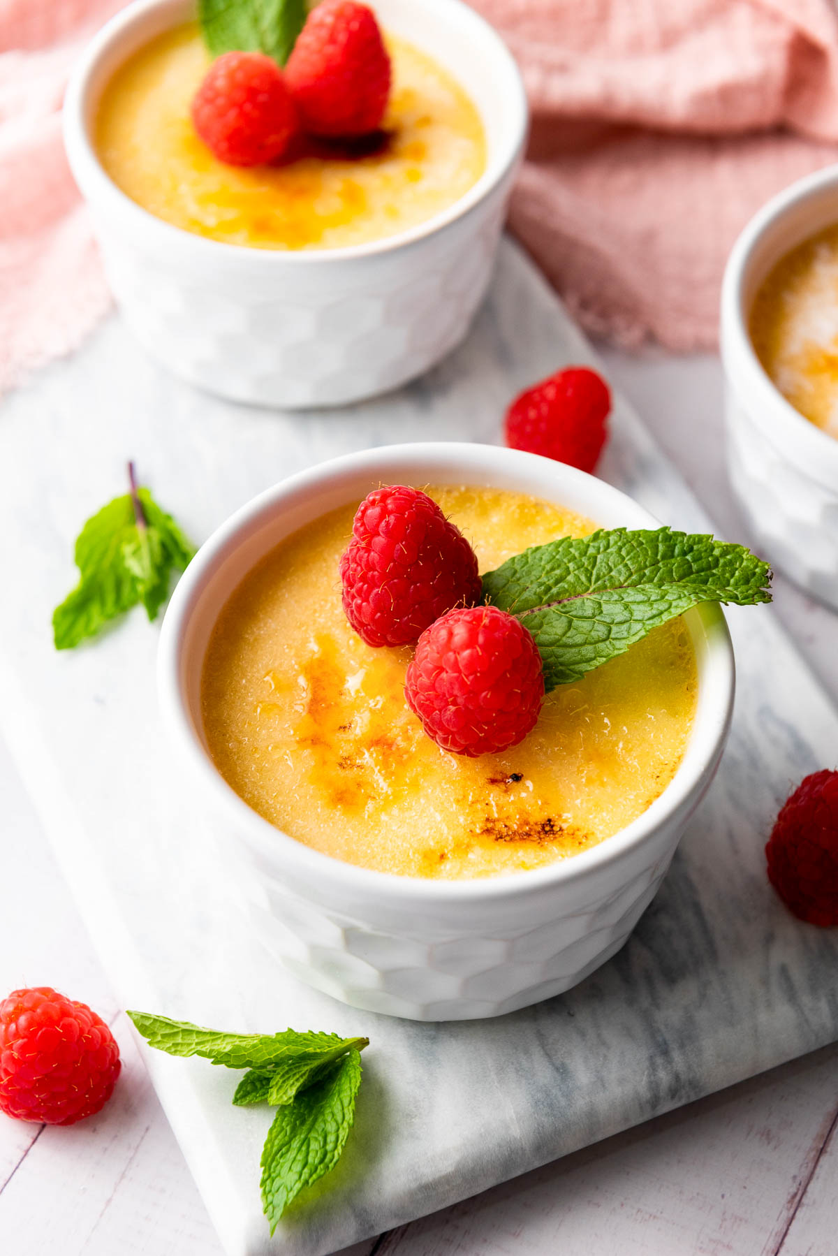 Two ramekins of crème brûlée, a quintessential dessert from French cuisine, are elegantly topped with raspberries and mint leaves, sitting gracefully on a marble surface.