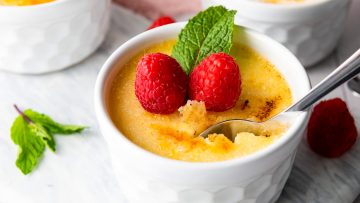 A bowl of silky crème brûlée is elegantly topped with two raspberries and a mint leaf, as a spoon takes an inviting bite.