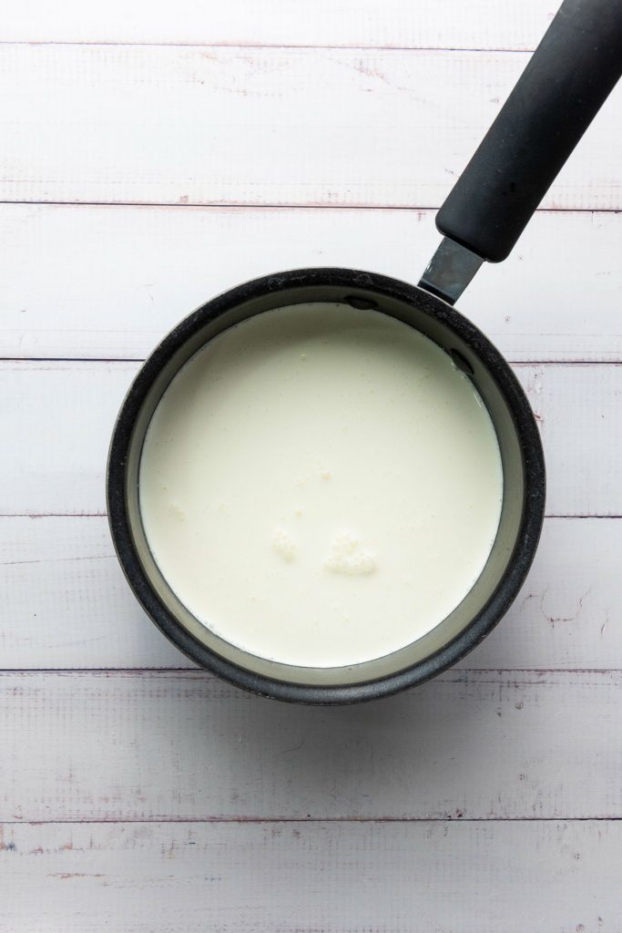 A black saucepan filled with a creamy white liquid, reminiscent of the first steps in crafting crème brûlée, sits elegantly on a white wooden surface.