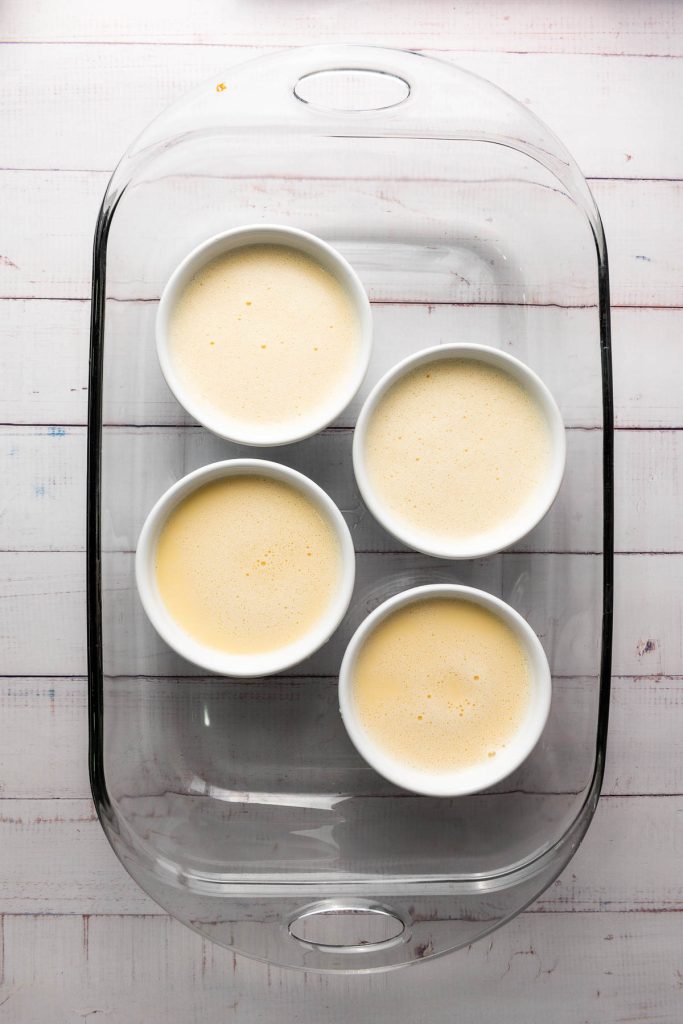 Four white ramekins filled with creamy Crème Brûlée custard are elegantly arranged in a clear glass baking dish on a light wooden surface, ready to delight any dessert enthusiast.
