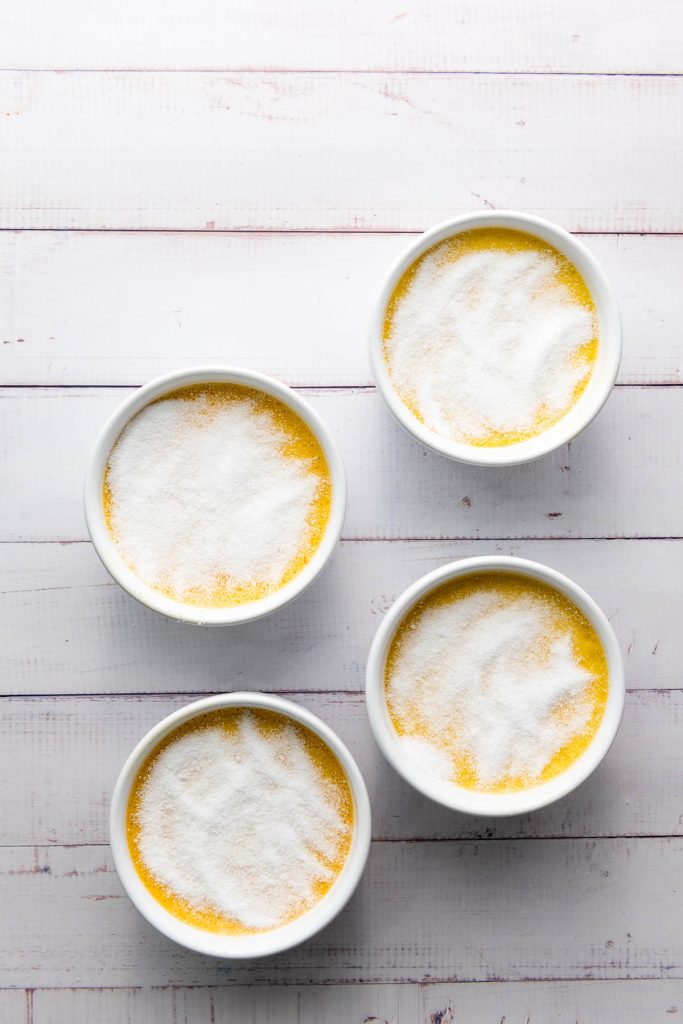 Four bowls of delicious crème brûlée topped with caramelized sugar rest invitingly on a white wooden surface.
