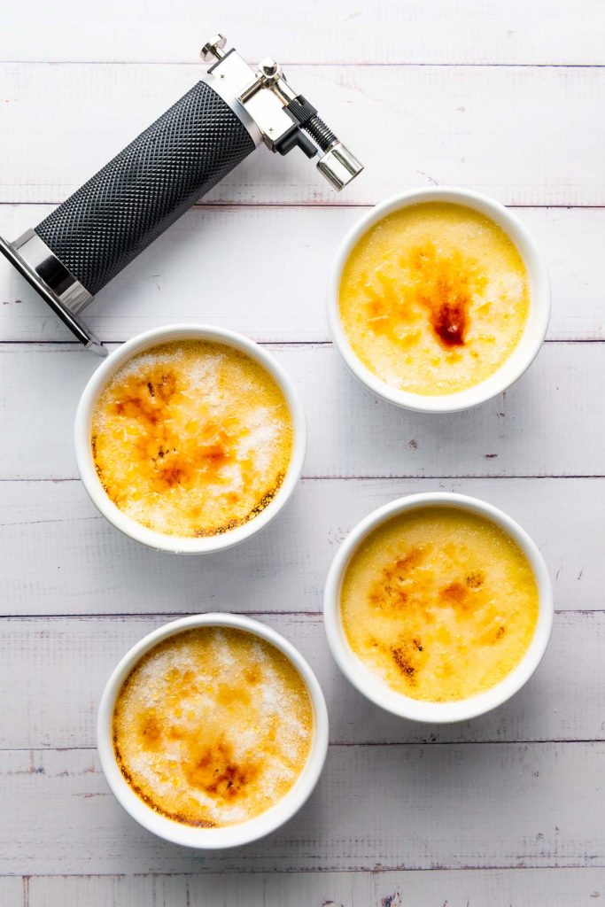 Four bowls of luscious crème brûlée with caramelized tops are artfully arranged on a white wooden surface next to a sleek black kitchen torch.