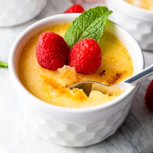 A small white ramekin of delicious crème brûlée is adorned with two raspberries and a mint leaf, while a spoon artfully breaks the caramelized sugar crust.