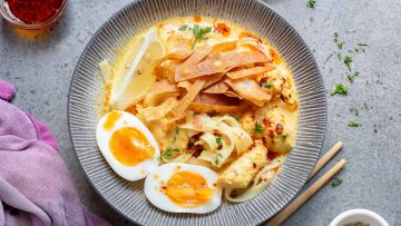A bowl of creamy soup with sliced boiled eggs, crispy tortilla strips, lemon wedges, and herbs, served on a gray plate with a pink cloth and chopsticks on the side.