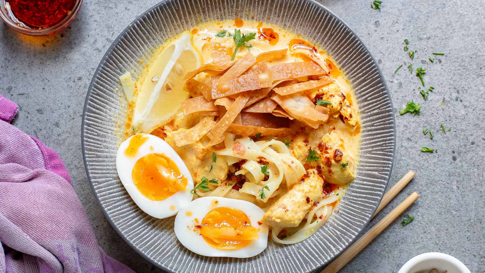 A gray bowl filled with a creamy noodle soup topped with sliced boiled eggs, crispy fried noodles, lemon wedges, and herbs. A napkin, chopsticks, and a small jar of spices are nearby on the gray surface.