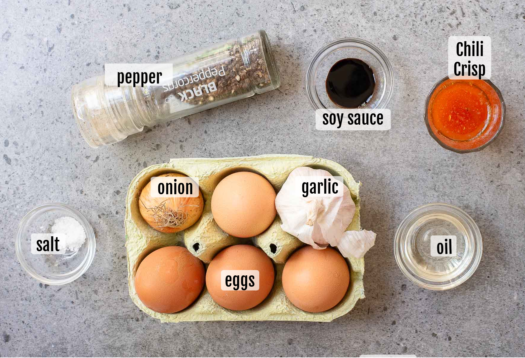 Ingredients for chili crisp fried eggs, including eggs, garlic, onion, salt, pepper, soy sauce, chili crisp, and oil arranged on a gray countertop.