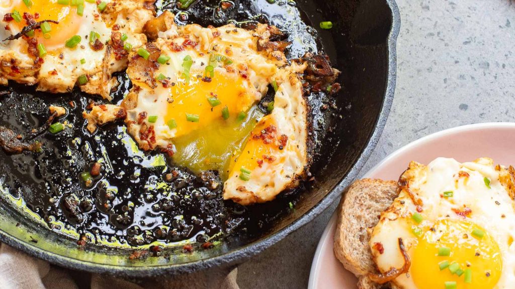 Chili Crisp Fried Eggs with herbs in a cast iron skillet sit invitingly next to a plate with egg-topped toast.