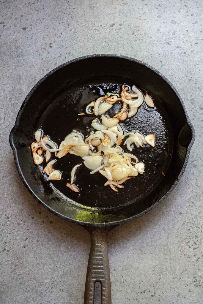 Sliced onions and garlic are being sautéed alongside chili crisp in a cast iron skillet on a gray surface.