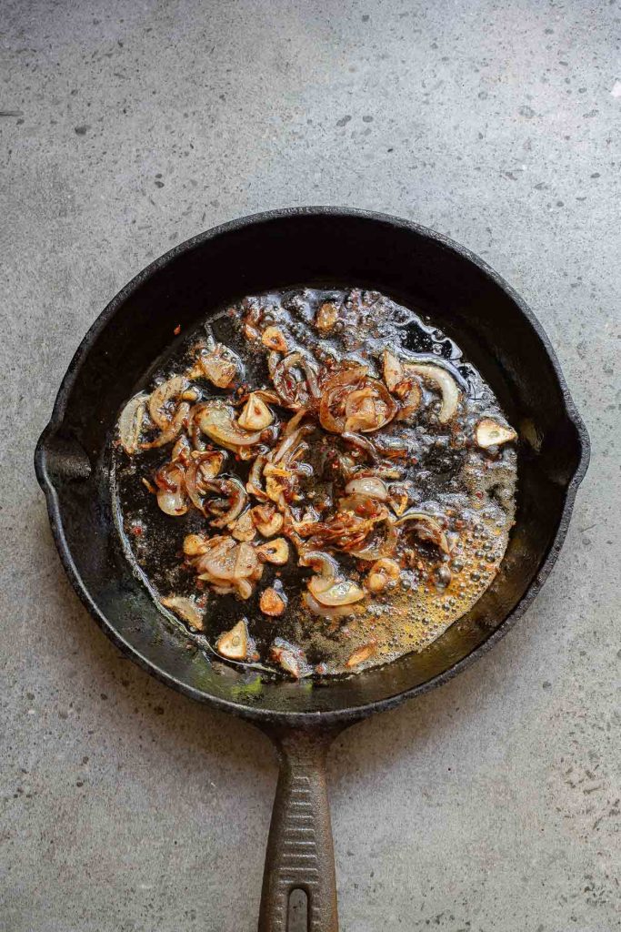 A cast iron skillet with sautéed onions, garlic, and a hint of chili crisp rests on a gray countertop.