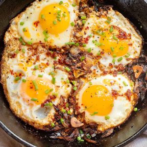 Four sunny-side-up eggs sizzle in a skillet, garnished with chopped chives and a hint of chili crisp for an extra kick.