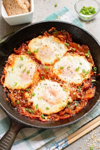 Four fried eggs on a bed of seasoned vegetables in a cast iron skillet, garnished with chopped green onions and sesame seeds, set on a checkered cloth with chopsticks nearby.