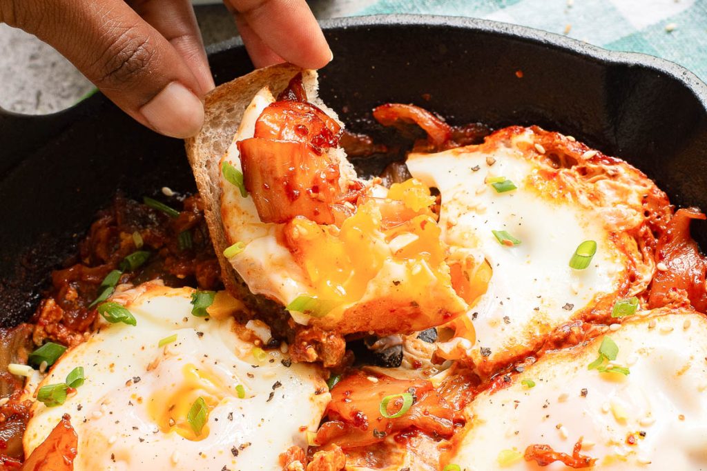 A hand holds a piece of bread topped with a bite of eggs and sauce from a skillet dish, garnished with green onions.