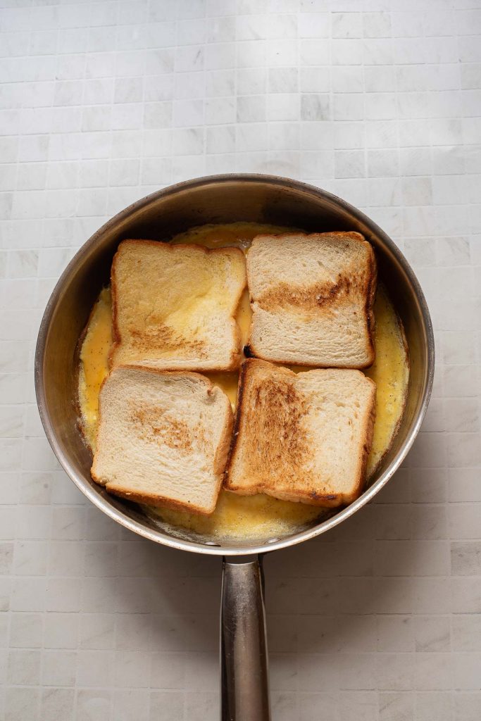 Four slices of toasted bread sit atop a layer of cooked eggs in a frying pan, creating a delicious one-pan egg sandwich on a tiled surface.