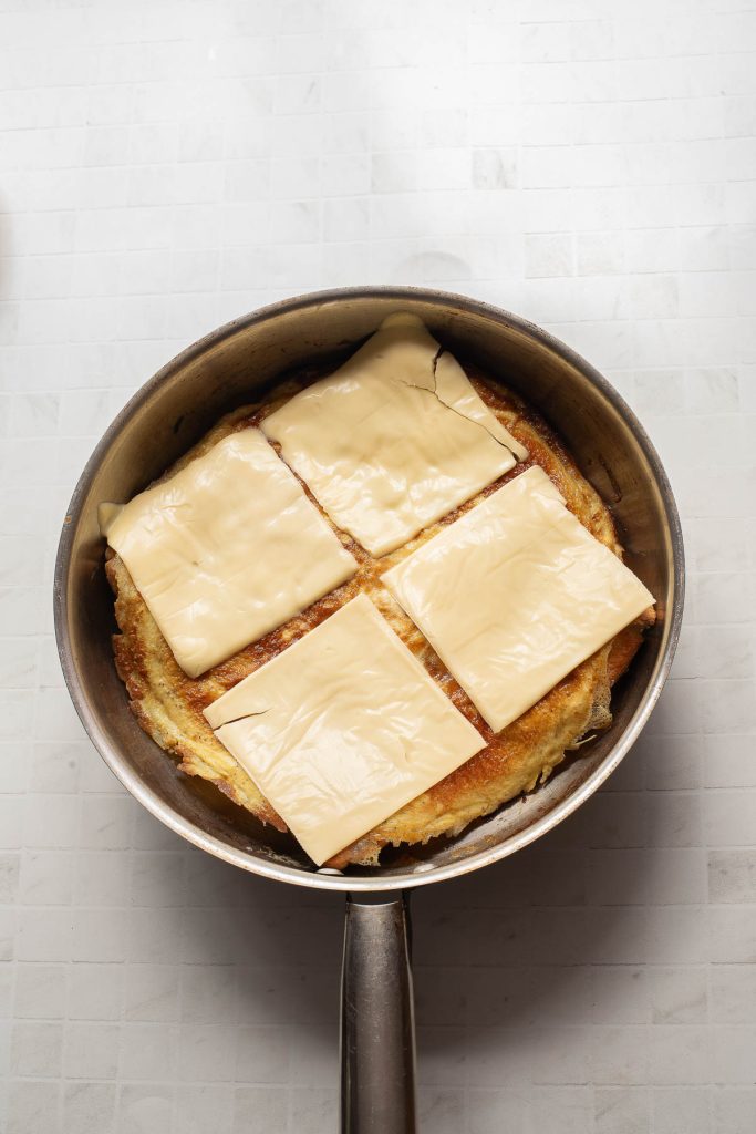 A frying pan containing a large piece of bread topped with four slices of processed cheese, perfect for an effortless one-pan breakfast, set on a light-colored tiled surface.