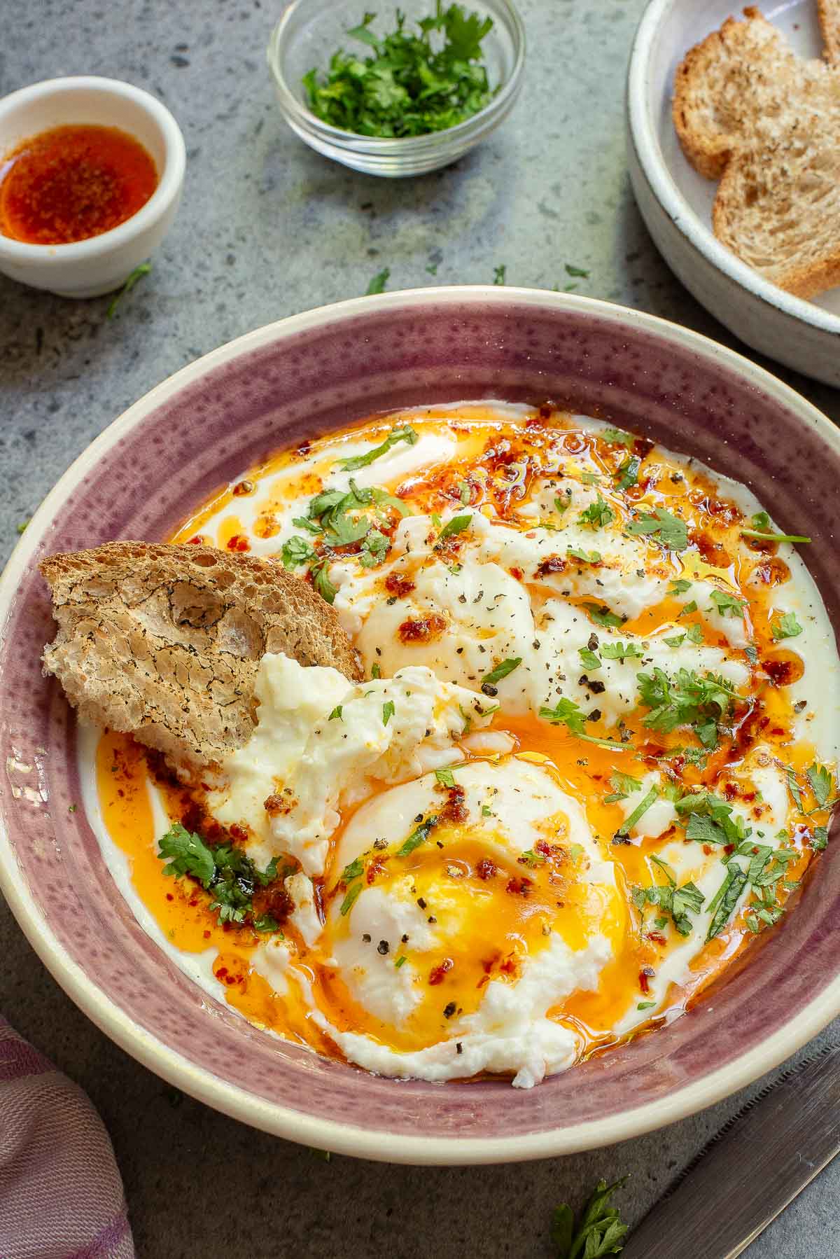 A bowl of Turkish Eggs: poached eggs topped with chili butter and chopped herbs, served with a piece of bread on the side; toast, sauce, and herbs are in the background.