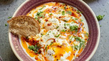 A bowl of Turkish Eggs: poached eggs over garlic yogurt, topped with herbs and chili butter, served with a slice of bread on the side.