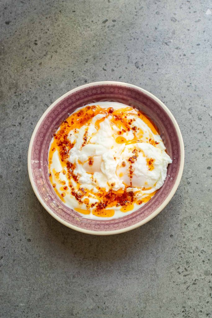 A bowl of Turkish Eggs—poached eggs in garlic yogurt—topped with rich chili butter and red pepper flakes, sits on a gray stone surface.