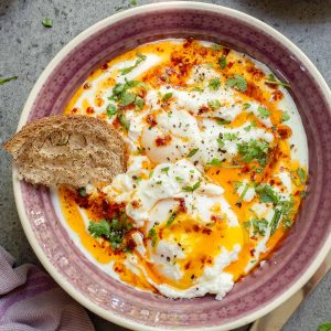A bowl of Turkish Eggs with poached eggs nestled in garlic yogurt, topped with chili butter, herbs, and black pepper, served alongside a slice of crusty bread.