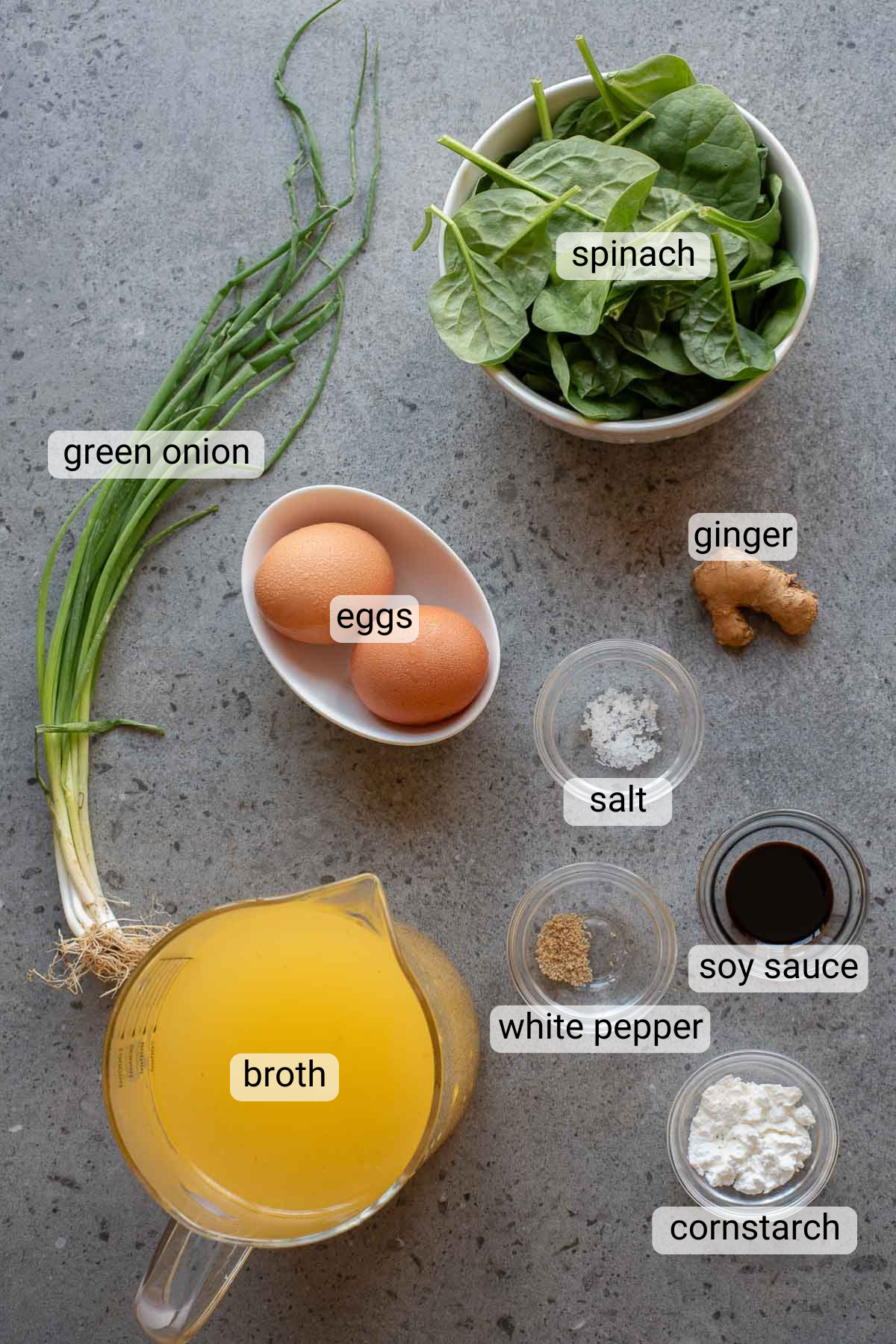 Ingredients for Egg Drop Soup on a gray surface: green onion, eggs, spinach, ginger, salt, white pepper, soy sauce, cornstarch, and a pitcher of broth are neatly labeled for easy preparation.