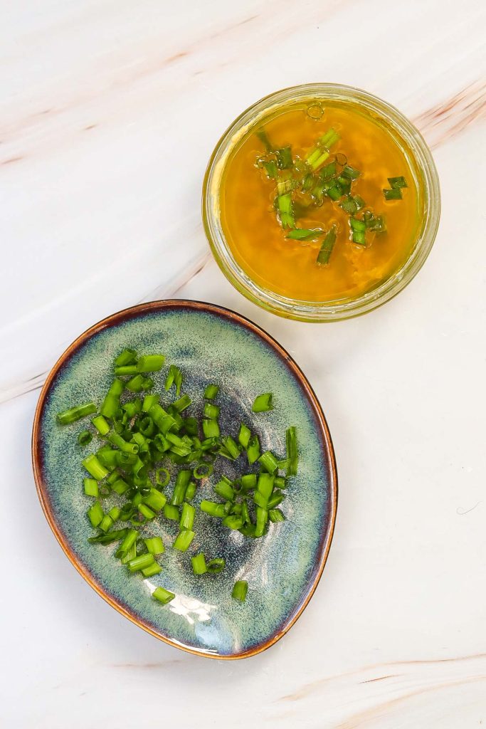 A blue plate with chopped green onions sits next to a small glass bowl of yellow liquid garnished with green onions, perfect for pairing with soft-boiled eggs on a white surface.