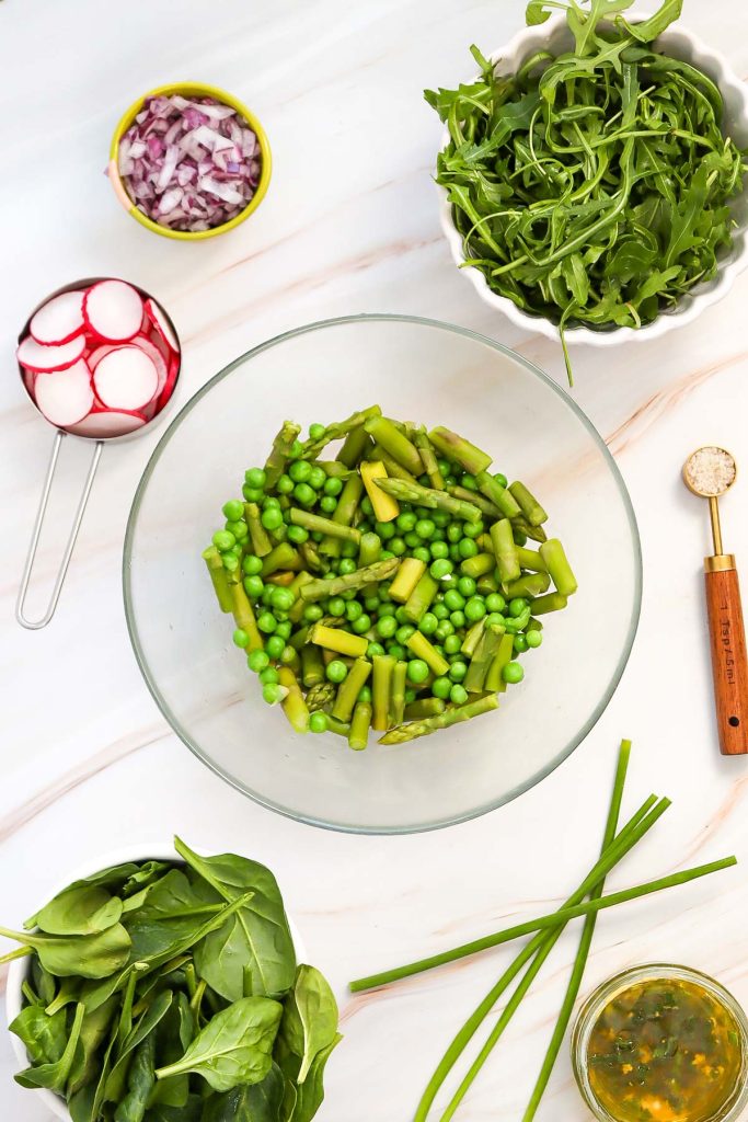 A bowl of chopped asparagus and peas forms the base of this vibrant asparagus salad, surrounded by arugula, spinach, sliced radishes, diced red onions, chives, and a small bowl of vinaigrette on a white surface.