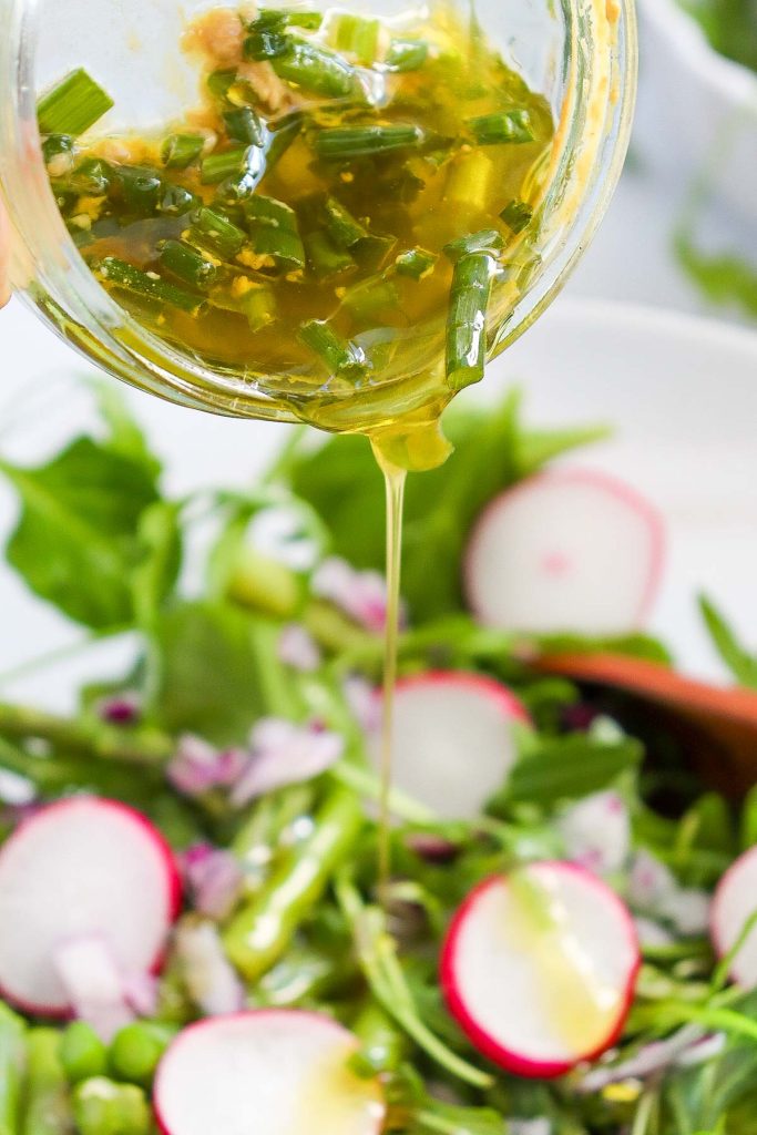 A jar of vinaigrette dressing is being poured over a fresh asparagus salad with sliced radishes, greens, chopped red onions, and soft-boiled eggs.