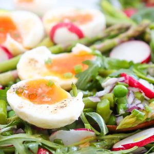 A close-up of a vibrant pea salad with halved soft-boiled eggs, tender asparagus, fresh peas, radishes, arugula, and chopped chives.