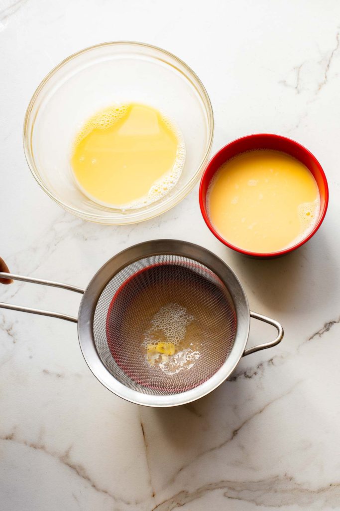 A fine-mesh strainer with some residue sits over a bowl, beside two bowls containing whisked eggs on a white marble surface—ingredients ready for a classic Chinese Steamed Egg Recipe.