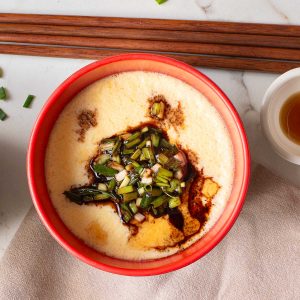 A red bowl filled with Chinese Steamed Egg custard, topped with chopped green onions and soy sauce, sits on a beige cloth next to chopsticks—a comforting Chinese recipe perfect for any meal.
