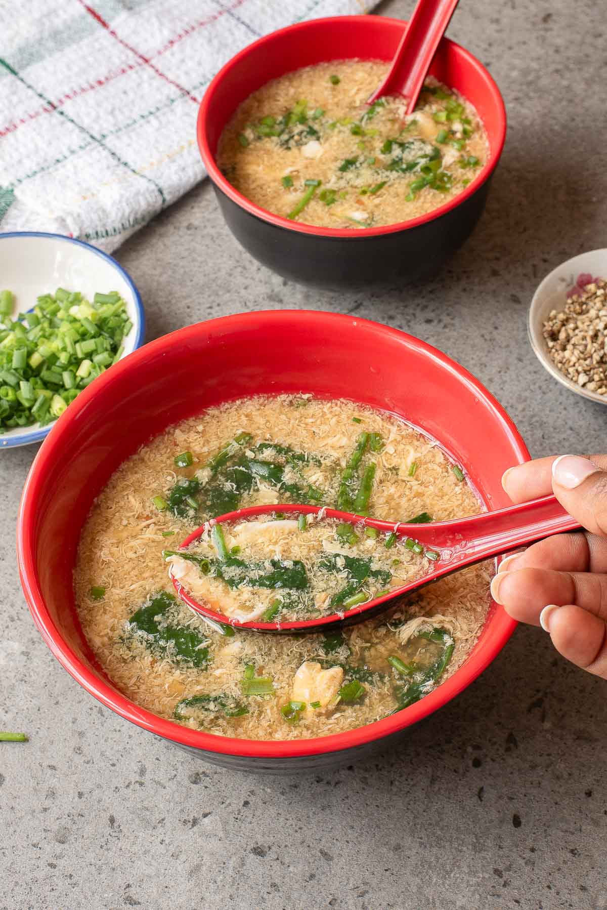 Two red bowls of Egg Drop Soup with green onions and spinach, one being held with a red spoon, sit on a gray surface beside small dishes of chopped green onions and pepper.