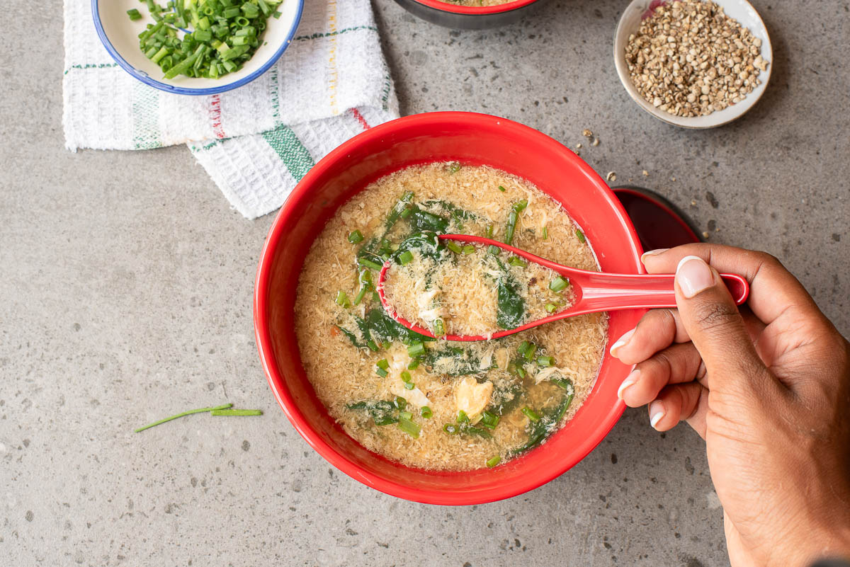 A hand scoops Egg Drop Soup with greens and egg from a red bowl, while chopped green onions and seasonings sit in small bowls nearby on a gray surface.