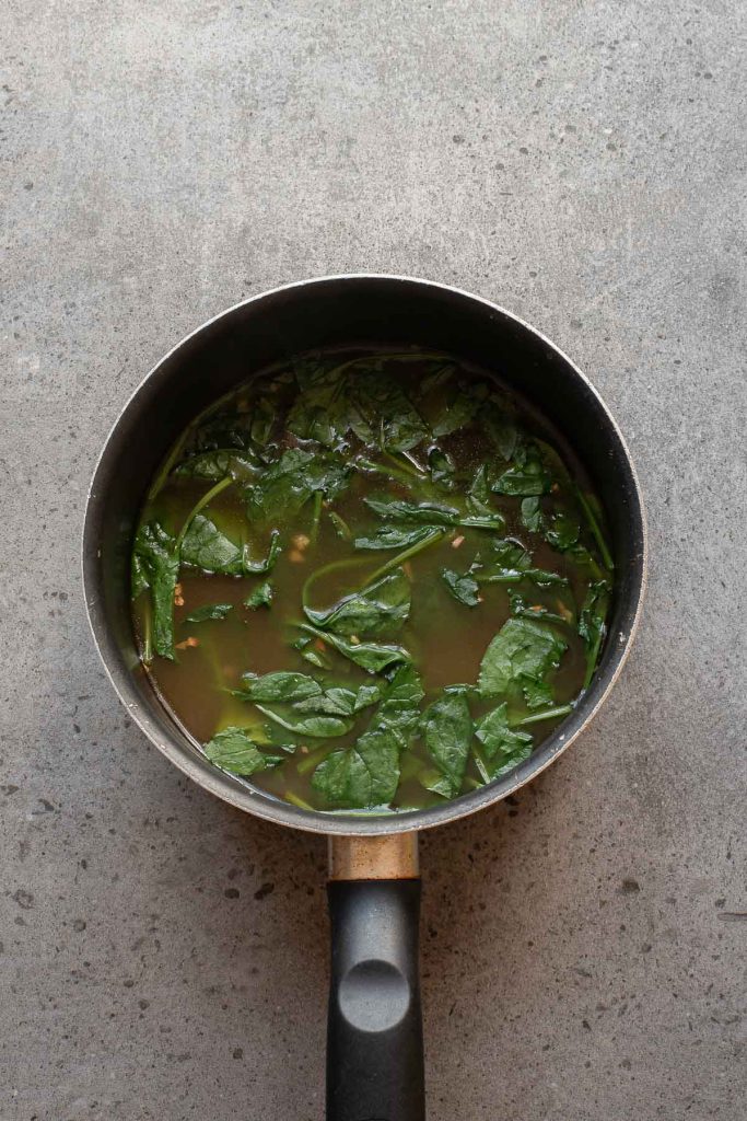 A saucepan filled with green leafy Egg Drop Soup sits on a gray textured surface, viewed from above.