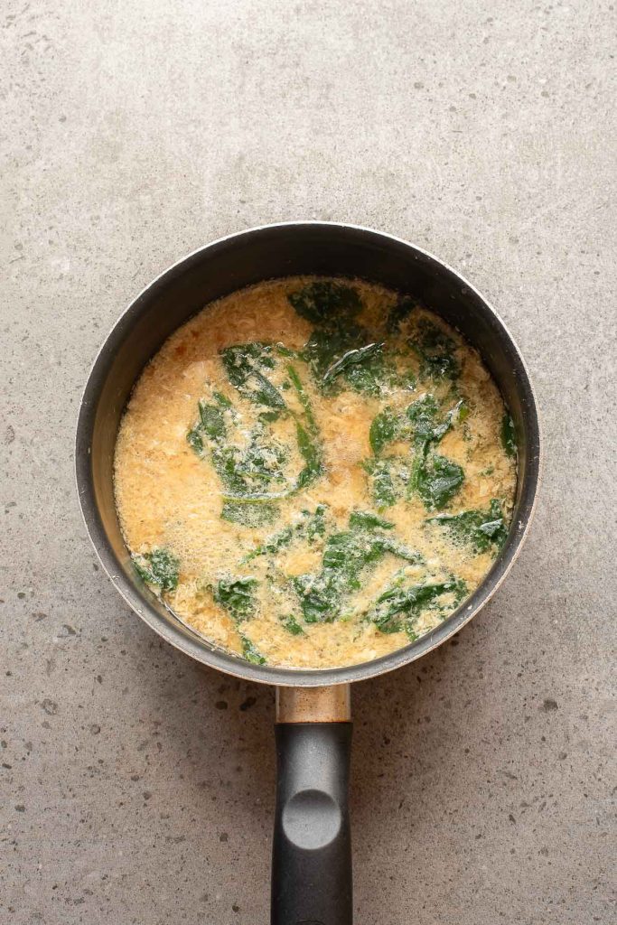 A saucepan filled with a creamy mixture containing wilted spinach, reminiscent of the comforting textures found in Egg Drop Soup, sits on a gray countertop.