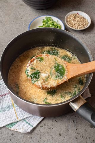 A pot of Egg Drop Soup with spinach is being stirred with a wooden spoon; nearby, small bowls hold chopped green onions and sesame seeds.
