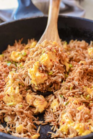 A wooden spoon stirring Spicy Egg Fried Rice with scrambled eggs and green onions in a black skillet.