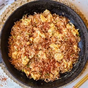 A black skillet filled with Spicy Egg Fried Rice, scrambled eggs, and sprinkled spices, placed on a woven mat with chopsticks nearby.