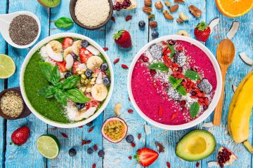 Two smoothie bowls with assorted fruit toppings sit on a blue wooden table, surrounded by fresh fruits, seeds, nuts, and a wooden spoon—a vibrant display of popular food trends in healthy eating.