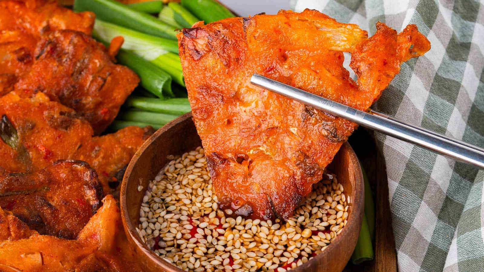 A piece of crispy, golden-orange Korean pancake held by metal chopsticks is being dipped into a bowl of sesame seeds and sauce. Green onions and a checkered cloth are in the background.