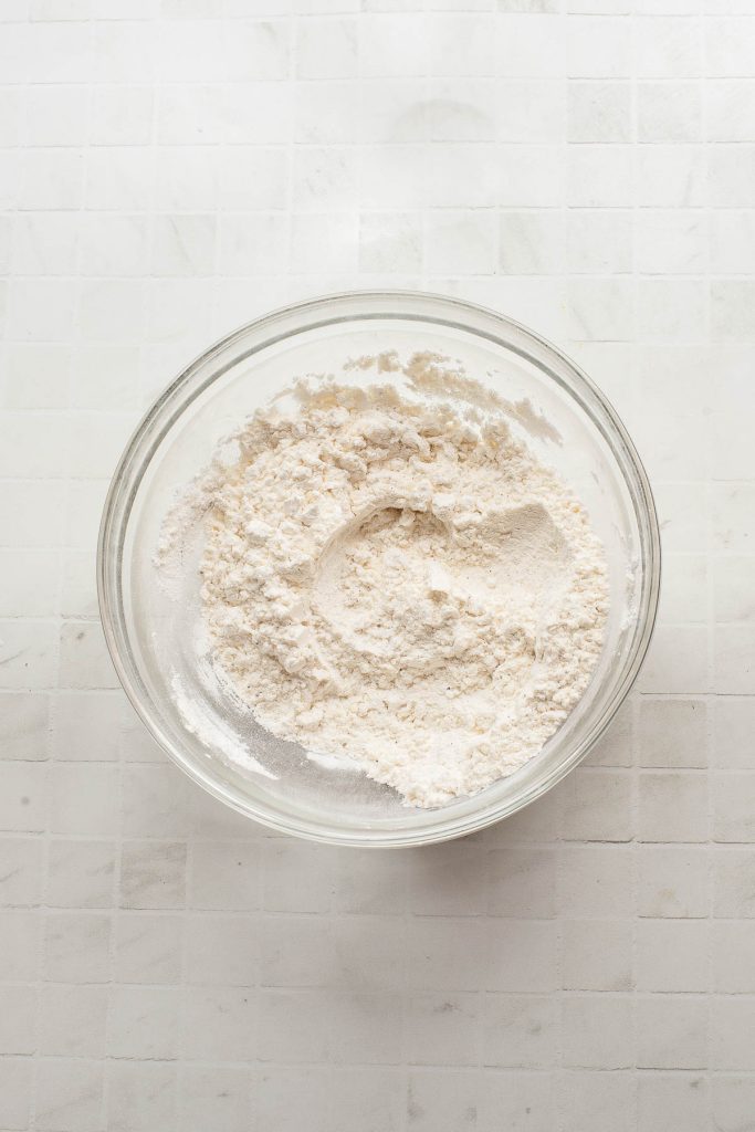 A glass bowl filled with flour mixture sits on a white tiled surface.