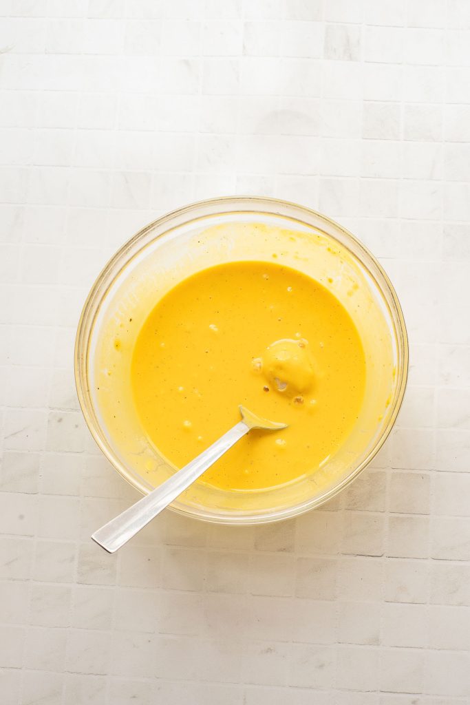A glass bowl filled with yellow batter and a metal spoon, placed on a white tiled surface.
