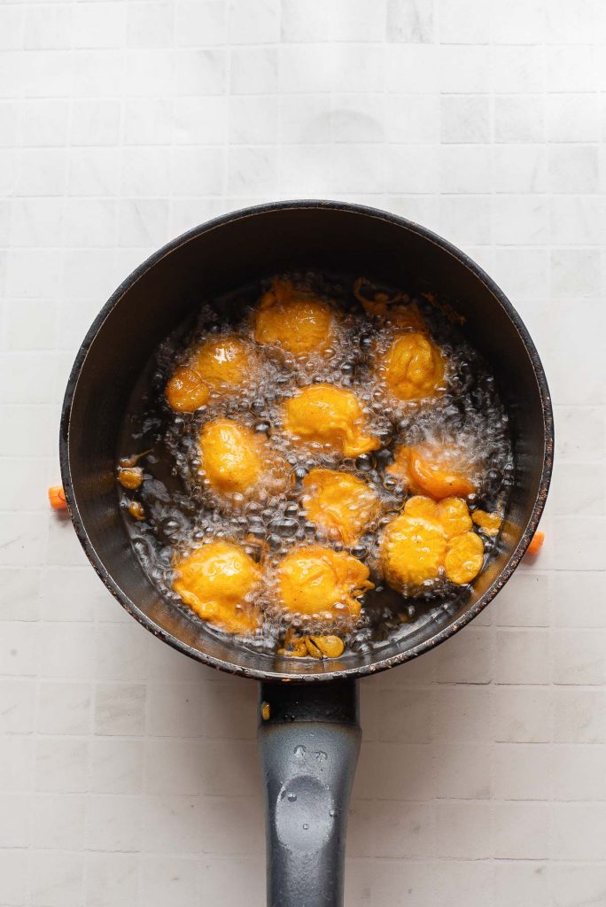 A black saucepan with several yellow dough balls frying in hot oil on a light tiled surface.