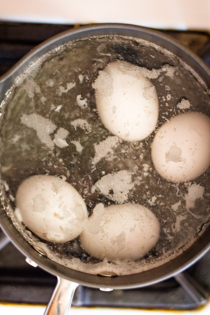 Four white eggs are boiling in a pot of water on a stovetop, with bubbles and some foam on the water’s surface.